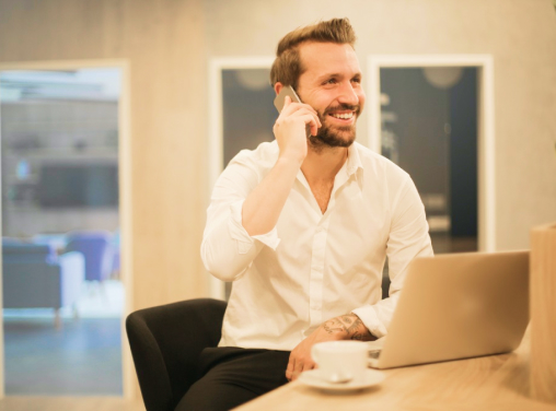 Homem sorrindo falando ao celular com um notebook e uma xícara de café a frente